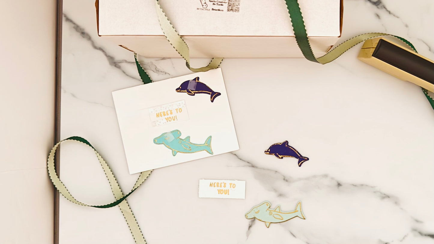 A tactile greeting card featuring a light teal shark at the top and a dark blue dolphin at the bottom, both with gold embossed outlines. The sea creatures appear to pop off the card. Centered between them is a white panel with the raised, gold embossed greeting: "Here’s To You." The overall card offers a cheerful, playful ocean theme with tactile details.