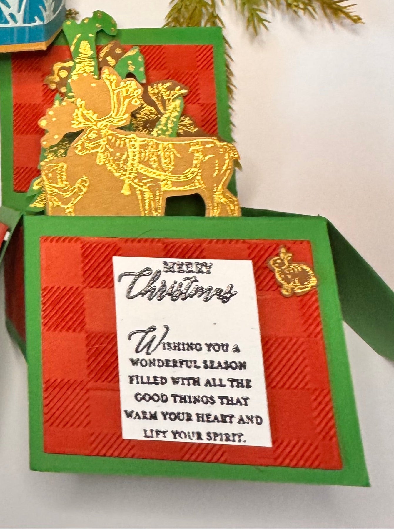 Gift set includes wax seal red background and deep green pine cone and branches that are 3 dimensional
Below it, a teal green and teal blue decorated box popup card base, with green teal leaves and cardinals (green teal) sticking out of the base of the card.
A red embossed like flannel and green background pre-made popup card with gold accents on brown reindeer, child and trees. On a white background with black writing is the greeting: