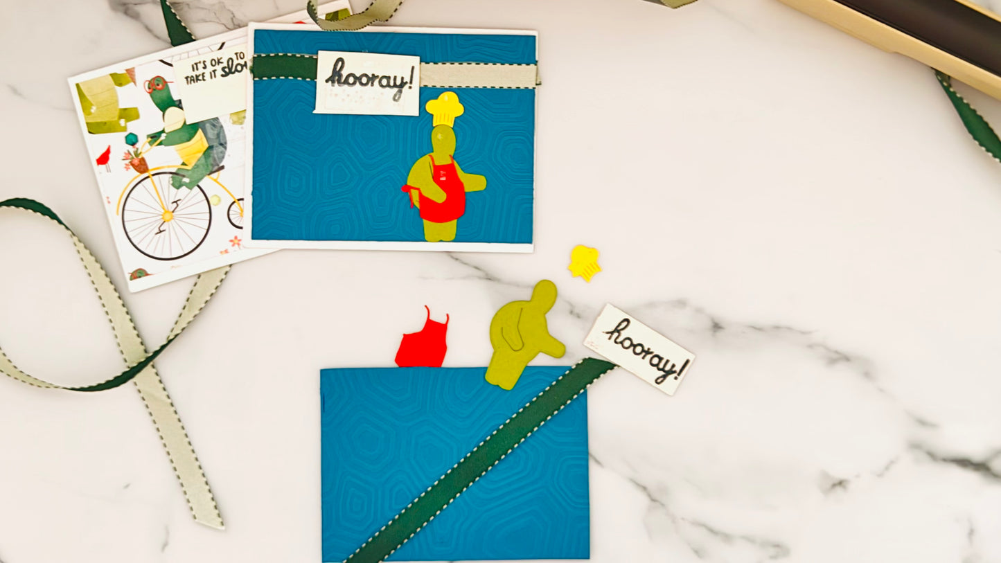 Handmade tactile card on deep blue embossed paper with raised hexagon pattern resembling a turtle shell. Features a smiling turtle wearing a chef’s hat and red apron, holding a spatula, with large tactile text "Hooray!" on a cream banner. Card is positioned on a white marble surface.