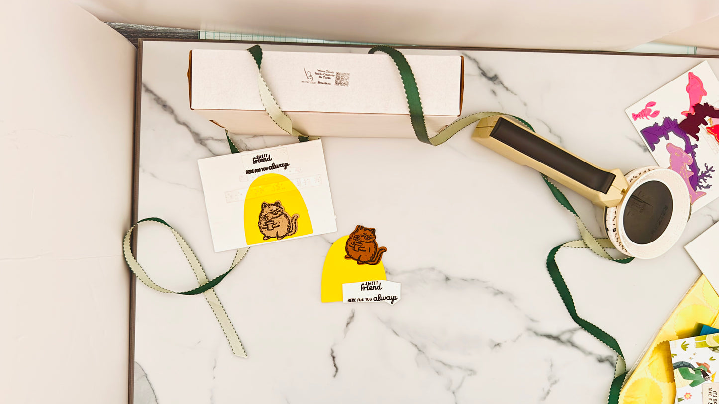 A small light brown fluffy cat holds a embossed cutesy flower in a flowerpot, standing in front of a yellow-orange half-circle background. The Tactile Treasures box is on its side, with a ribbon swirling across the photo and around a handheld broiler positioned at the top right.