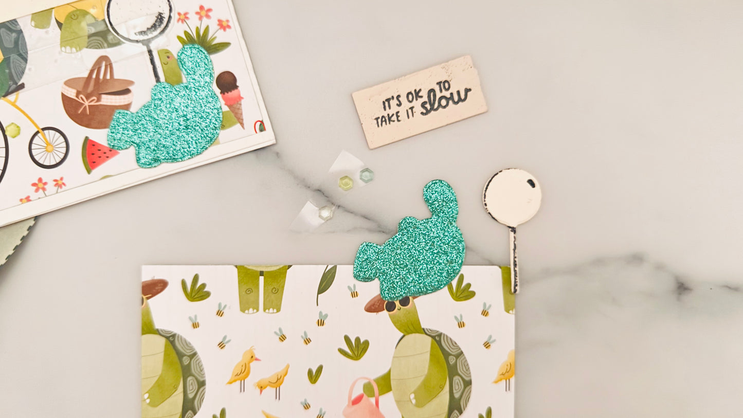 Close-up of a handmade tactile card kit in progress on a white marble surface. The main card design shows a turtle-themed picnic scene with illustrated turtles wearing hats and sunglasses, holding picnic baskets, surrounded by birds, bees, flowers, watermelon, ice cream, and a bicycle. Two glittery, textured turquoise turtle shapes are included as tactile elements—one attached to a card in the upper left corner, the other lying loose on the illustrated background. A small cream-colored plaque with raised bl