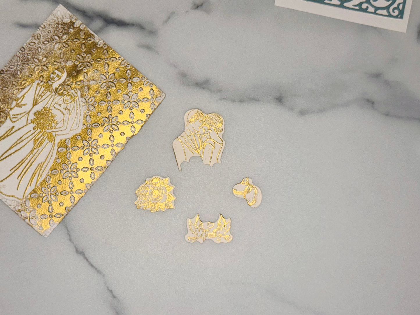 Accent pieces to add to wedding card: a small white cutout with gold line art of two interlocking wedding rings a small white cutout with gold line art of two dove birds, a piece of ribbon held between there beaks a small white cutout with gold line art of a rose (flower head with layered petals) a medium white cutout with gold line art of a couple in an embrace (upper bodies close together)