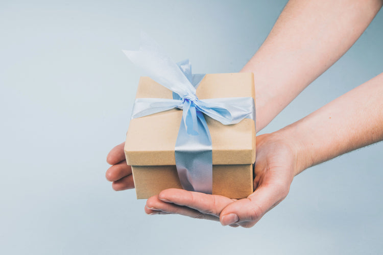 hands holding a small box that holds Be Tactile's card kits. Box is brown with a lid and a light blue ribbon around the box tied in a bow.