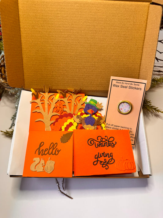 This is an open brown cardboard box containing autumn-themed stationery items. Inside the box, there one orange folded cards in the front. The card on the left says "hello" with images of a squirrel and an acorn. The card says "Thanks Giving" with a pumpkin illustration. Behind the cards, there are several cut-out paper decorations with fall designs, including trees and leaves in orange, yellow, and brown. On the right side, shows "Made By Hand Be Tactile Wax Seal Stickers, to place on the back of envelope.