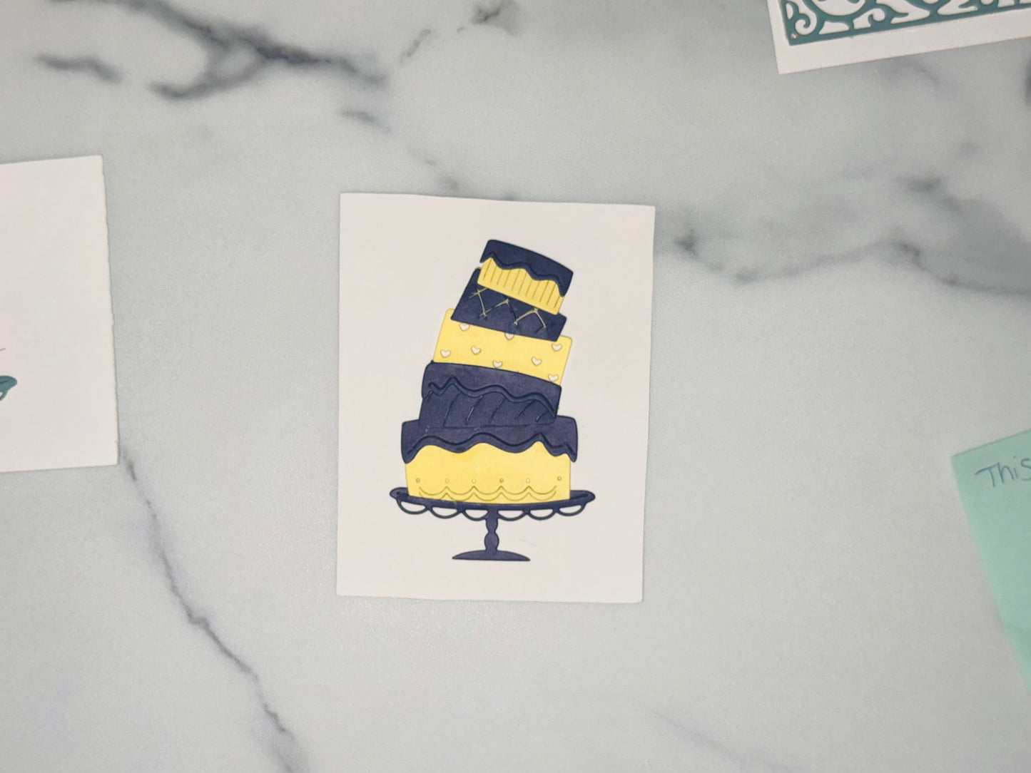 Bottom layer: Yellow cake with small dot accents and a scalloped, looped pattern near the bottom edge; dark navy icing drips in a wavy line around the top. Dark navy cake stand with a scalloped rim and a pedestal base. Middle layer: Dark navy layer with an embossed-looking wavy pattern across it; topped with another dark navy wavy/drippy icing band. Upper layer: Yellow layer with small white heart shapes scattered across it; a thin dark navy band sits on top of this layer. Top layer: Dark navy top 