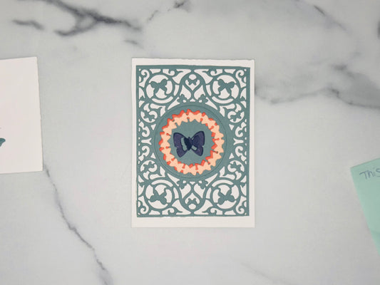 inner teal-gray butterfly panel that looks like a lacy cut-out is a symmetric filigree pattern made the corners are butterflies. In the center is a teal-gray large circle. Inside it is a ring of cut out butterflies overlapping one is a soft burnt orange shade and the other is a light peach pink outline along the outer edge, like a decorative ruffle. In the middle of the circle is a dark navy butterfly (like a ribbon bowtie) with a smaller butterfly in the center.