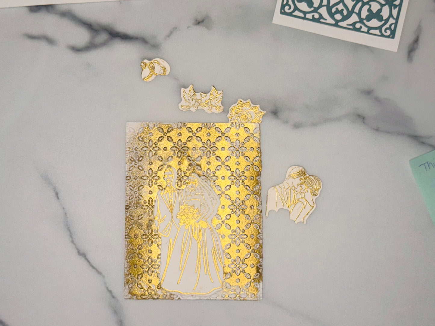 Gold background filled with a 3-D repeating pattern of small flower shapes, cutout with gold line art of a bride and groom standing together The groom is in a suit; the bride wears a long dress and a veil and holds a bouquet at waist level Accent pieces to add to wedding card: a small white cutout with gold line art of two interlocking wedding rings a small white cutout with gold line art of two dove birds, a piece of ribbon held between 