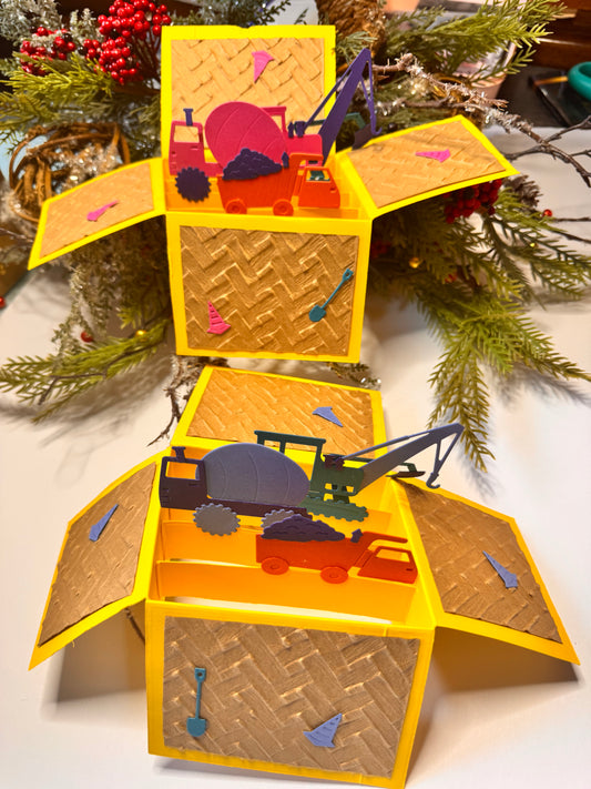 Handmade pop-up card box with two separate construction themes one pink shades and one blue shades. The box is mostly bright yellow, with a woven pattern on the panels, and each side drops open to reveal colorful paper cutouts of construction vehicles inside. Cut outs include shovel, cones, dump truck and dirt, cement truck, escalator or pulling/lifting type work truck with claw.