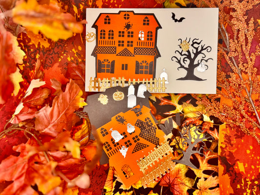 Orange paper cut-out haunted houses with black windows sit at the center, decorated with stickers of ghosts, bats, a spider, tombstones, a jack-o’-lantern, and a skull. Glittery texture orange paper fences frame the houses, while a stark black tree silhouette and pale cut-out branches add contrast. Scattered around are whimsical accents such as a floating ghost and a bat in flight, set against a vivid background of artificial autumn leaves in shades of orange, yellow, and red. The scene balances cozy fall.