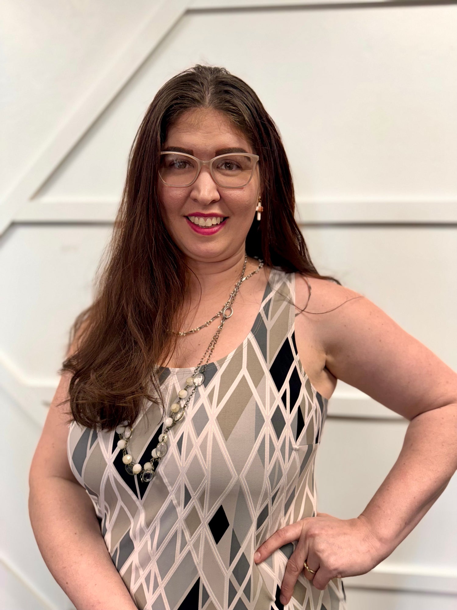 Jen is dressed in a sleeveless dress and has a V-shaped neckline.  The fabric pattern is made of long, narrow diamond and triangle shapes.  Colors in the pattern include white, light gray, darker gray, tan, and black.  The overall style is fitted at the top and looks like a smooth, soft material, giving a polished but comfortable appearance. Around her neck are layered necklaces, including one with pearls and another with small beads or links. One arm is bent with a hand on her hip, giving a confident pose.