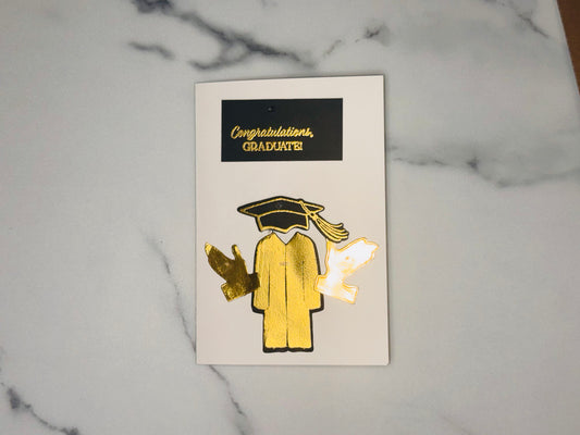 “Congratulations, GRADUATE!” Below that is a stylized graduate figure outlined in black. The graduation cap is black with gold trim and a gold tassel hanging to the right. The gown is metallic gold with a vertical seam line down the middle. On each side of the graduate are two separate hand-shaped cutouts: the left one is metallic gold, and the right one is white/light with a gold outline.