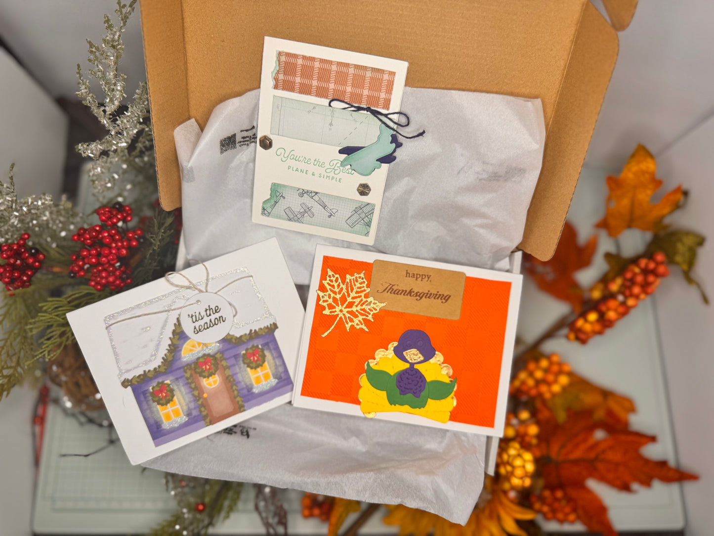 A collection of the October Subscription box hinting at Season of Gratitude: Honoring, Gathering, and Giving. Each box for October includes two Plane & Simple with plane accents to add and paper with plane themes a nod to support our Veterans. Two A Thanksgiving classic with embossed checkered paper, two maple leaves, and a detailed-layered turkey with four layers of feathers. The third card is A House Full of Cheer a simple yet cozy card carries the warmth of the holidays without leaning into traditions.