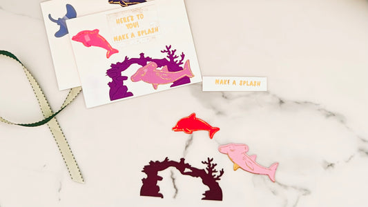 A tactile greeting card featuring a light pink shark playfully swimming across the top edge, and a dark pink dolphin gliding along the bottom, both with shimmering gold embossed outlines. A dark plum coral reef sits between the two sea creatures, adding rich texture and depth. Centered is a crisp white panel showcasing the raised, gold embossed greeting: "Here’s To You! Make a Splash." This cheerful ocean-themed card offers delightful tactile elements for all abilities.