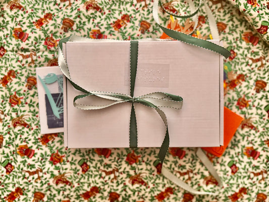 on vintage christmas fabric of wreathes, stockings and Christmas horns sits the white Tactile Treasures box with braille "Tactile Treasures" with a fancy two toned green ribbon tied neatly in a bow.