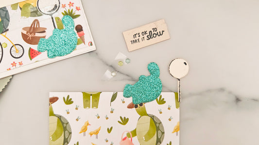 Close-up of a handmade tactile card kit in progress on a white marble surface. The main card design shows a turtle-themed picnic scene with illustrated turtles wearing hats and sunglasses, holding picnic baskets, surrounded by birds, bees, flowers, watermelon, ice cream, and a bicycle. Two glittery, textured turquoise turtle shapes are included as tactile elements—one attached to a card in the upper left corner, the other lying loose on the illustrated background. A small cream-colored plaque with raised bl